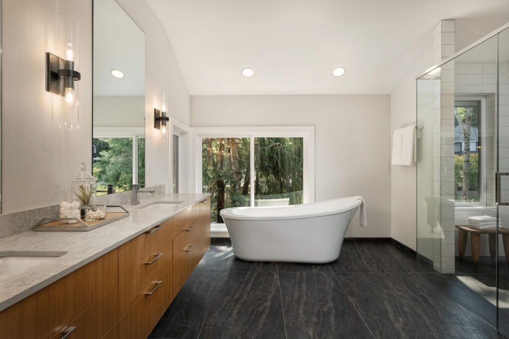 Luxury bathroom featuring a freestanding white bathtub centered in front of large windows with a forest view. The space includes a long wooden vanity with marble countertop, wall-mounted light fixtures, dark tile flooring, and a glass-enclosed shower on the right.