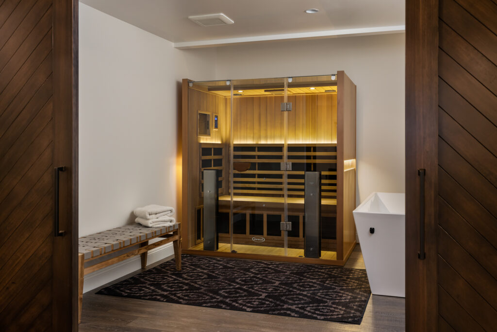 Sauna room with glass doors, wooden interior, and ambient lighting, situated in a clean, minimalist space. Features include a cushioned bench with folded towels on the left, a dark patterned rug in front, and a white container on the right, highlighting a relaxing wellness environment.